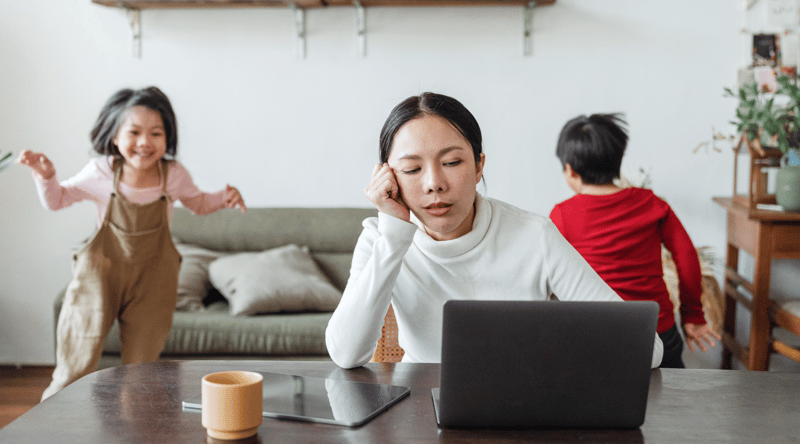 Frustrated woman who is considering quiet quitting on her laptop at home with her children playing in the background